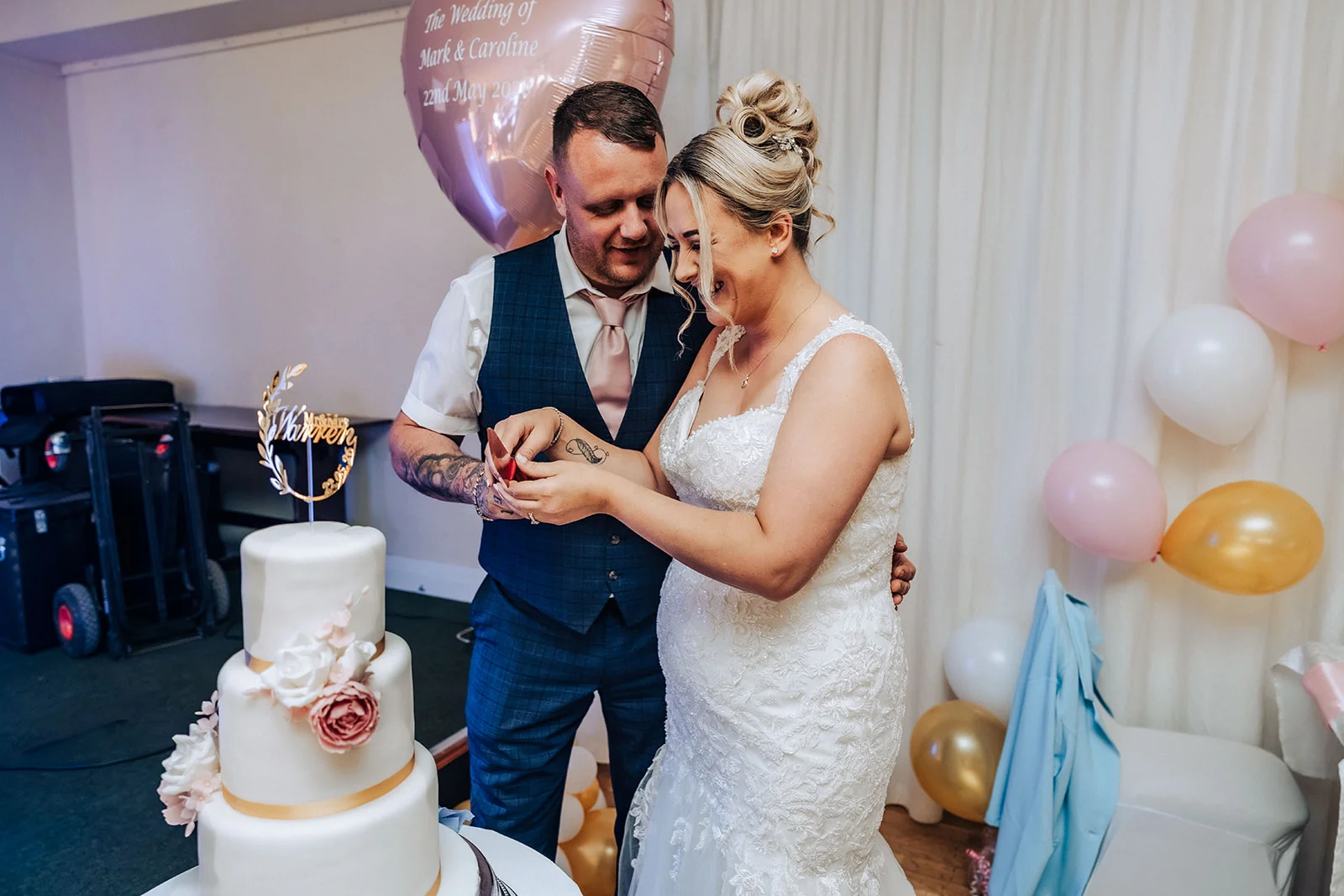 A joyful bride and groom laugh together while cutting their tiered white wedding cake decorated with pink and white floral accents.