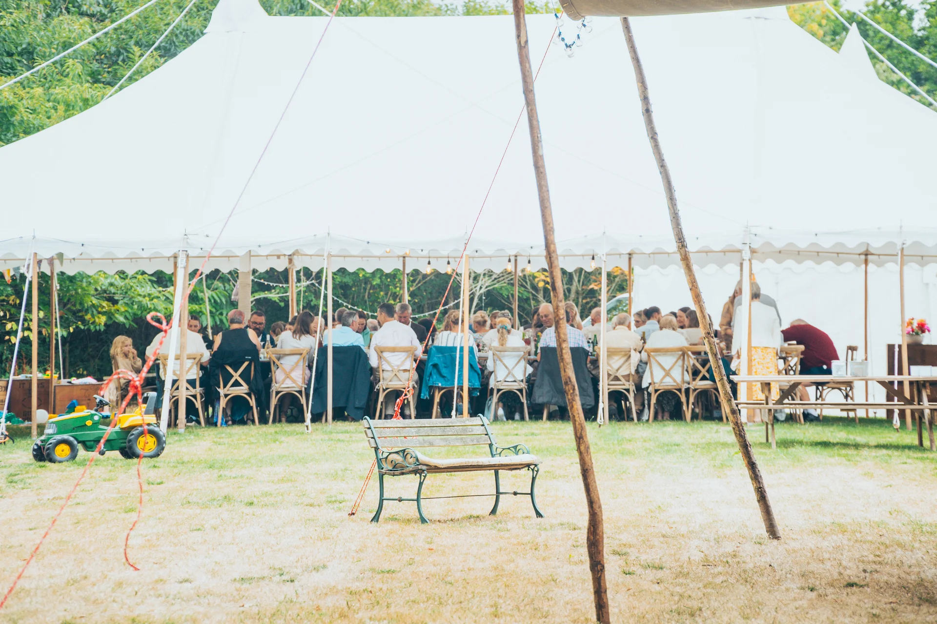 A wide outdoor shot of a large white sailcloth wedding marquee set up on a lawn, with guests seated at long banquet tables inside.
