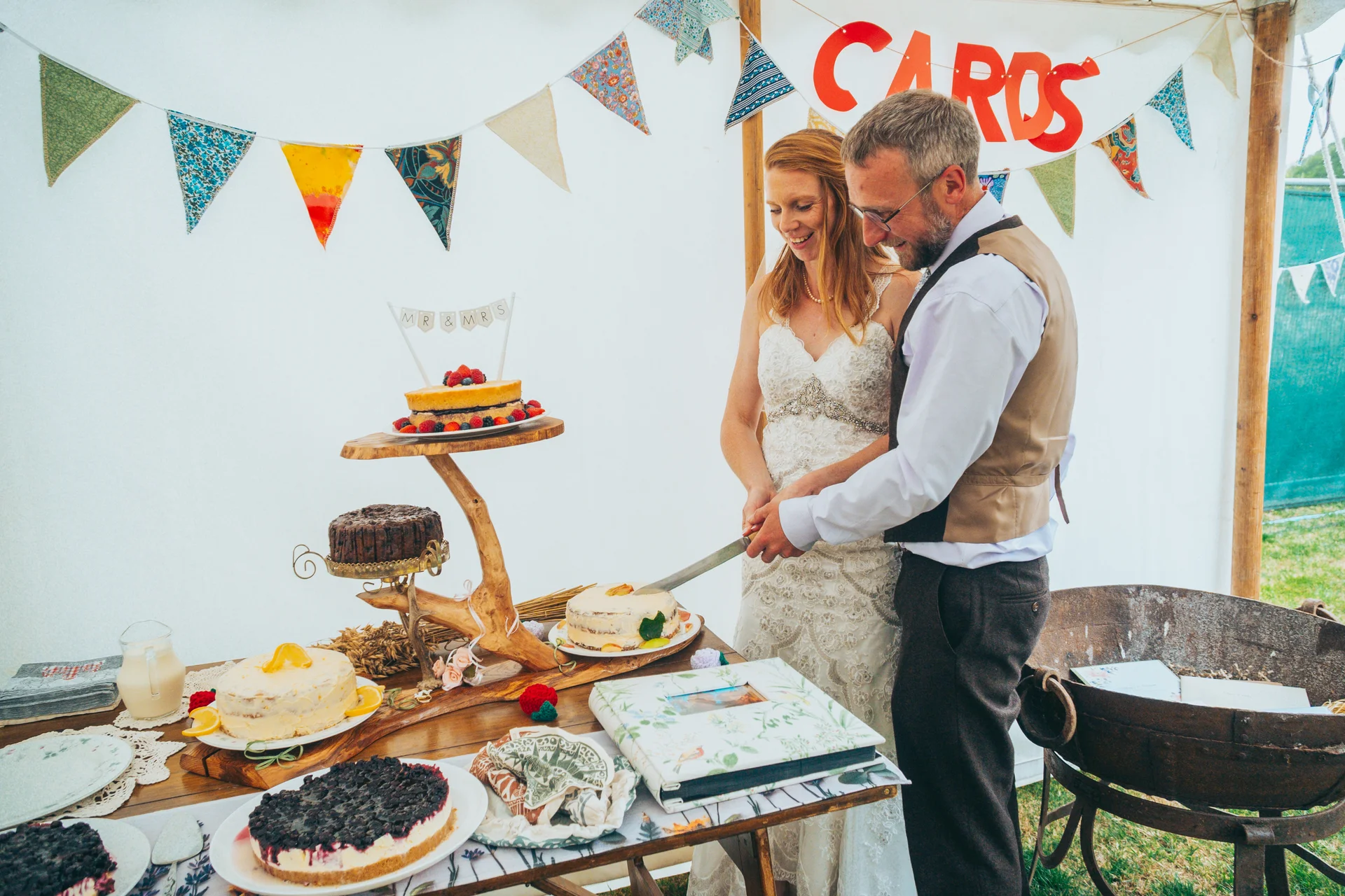 A bride and groom smile together inside a festival marquee as they cut into a rustic tiered wedding cake surrounded by various desserts.