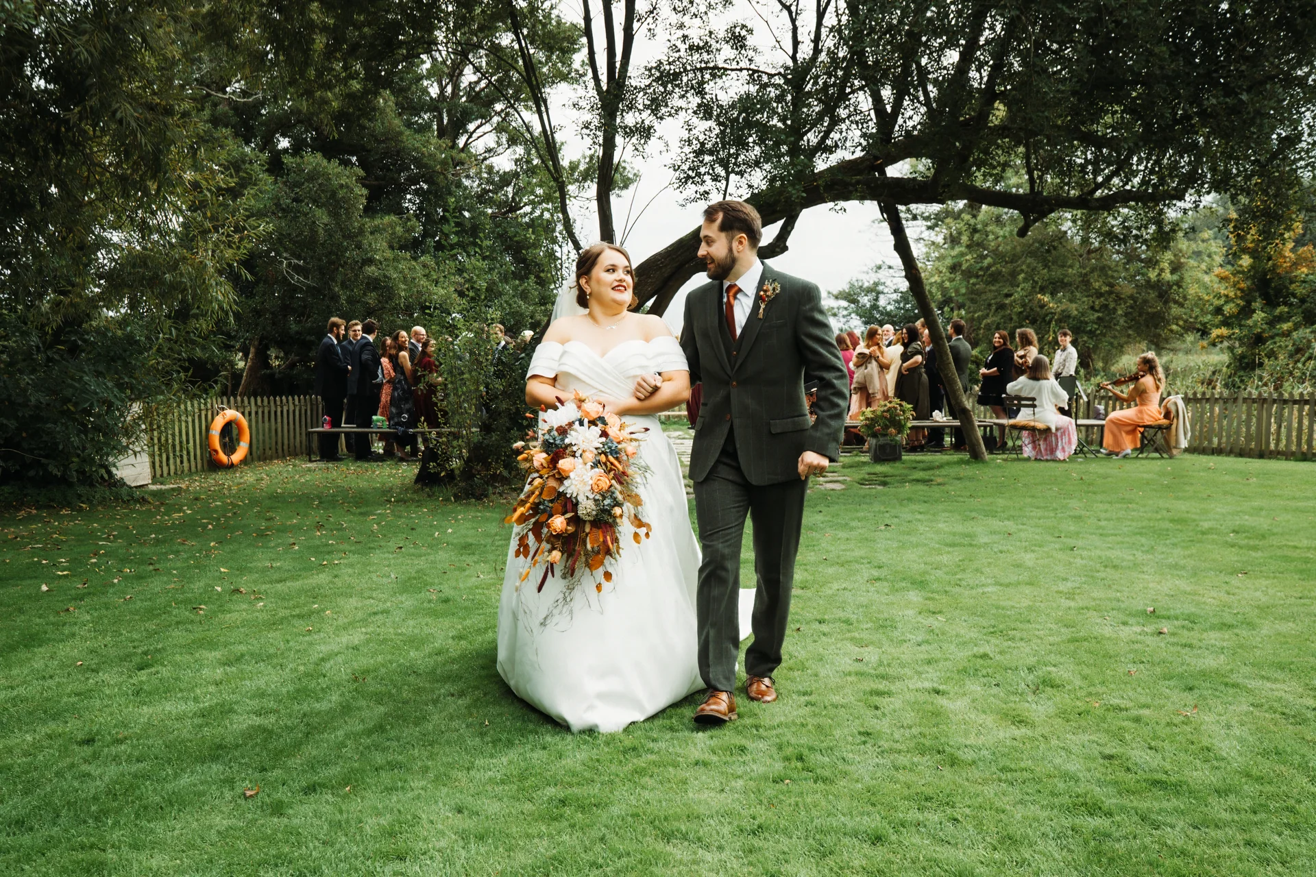 A newlywed couple walks across a lush green lawn holding hands, with the smiling bride carrying an oversized cascading autumn-toned bouquet and the groom wearing a dark green suit, while wedding guests watch from the background.