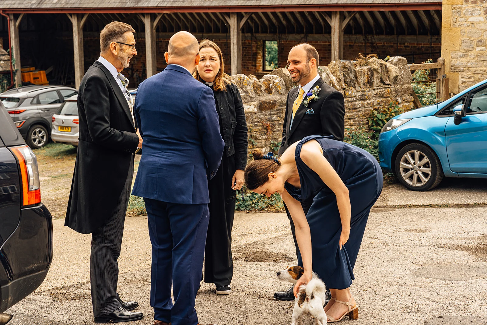 A wedding guest in a navy dress bends down to stroke a small terrier outside a rustic barn venue while other guests chat nearby.