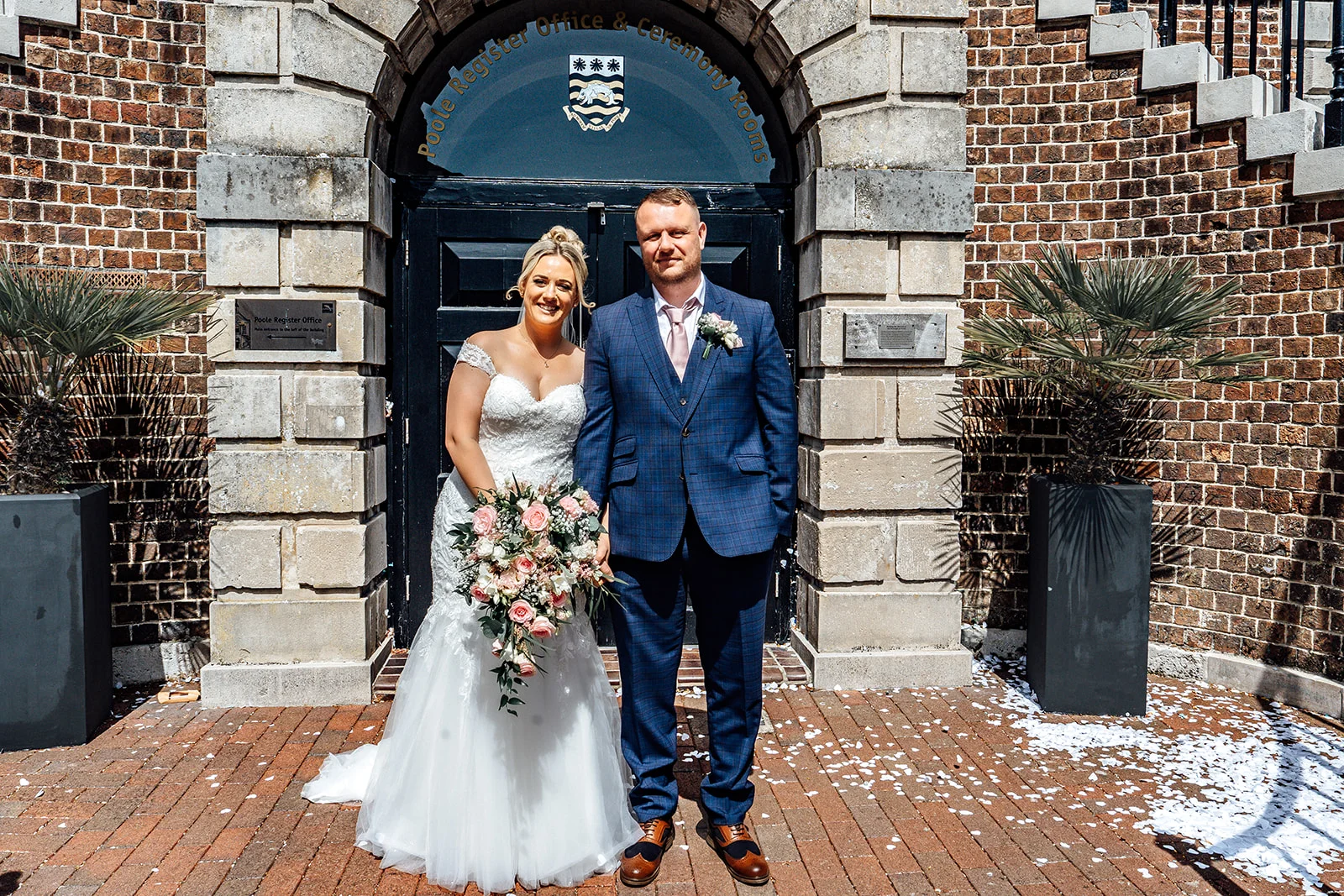 A newlywed couple poses outside a registry office entrance featuring a stone archway and brick walls, with the bride holding a cascading floral bouquet.
