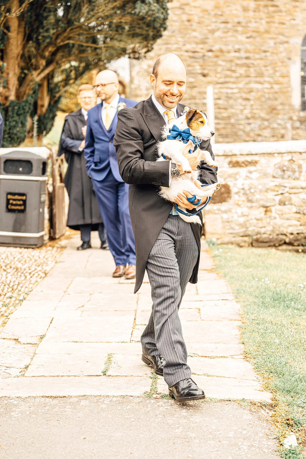 A smiling groom wearing a traditional morning suit walks along a stone path while carrying a small dog wearing a blue bow tie.