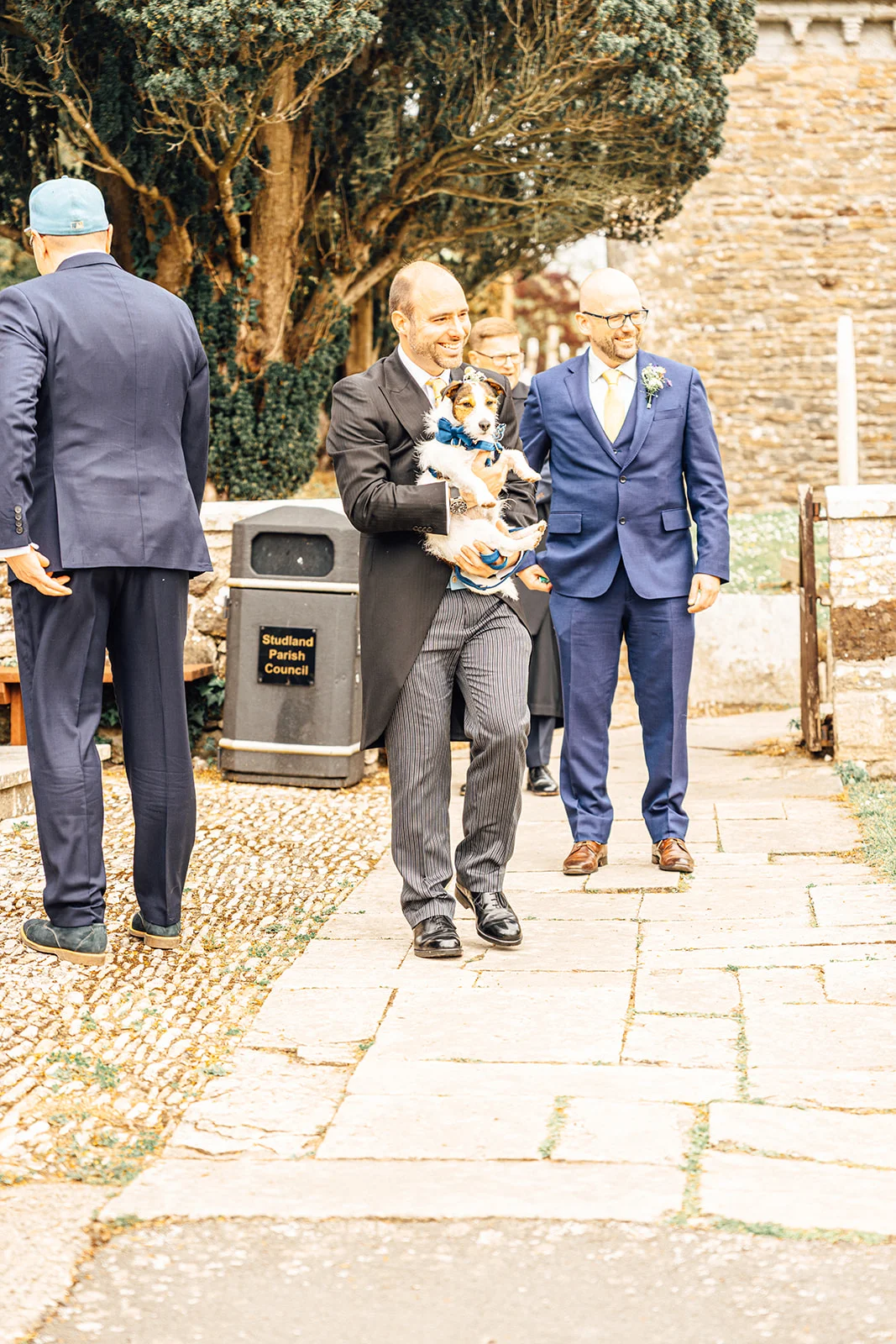 A smiling groom carrying a small dog wearing a blue bow tie walks along a stone pathway outside a church, accompanied by groomsmen.