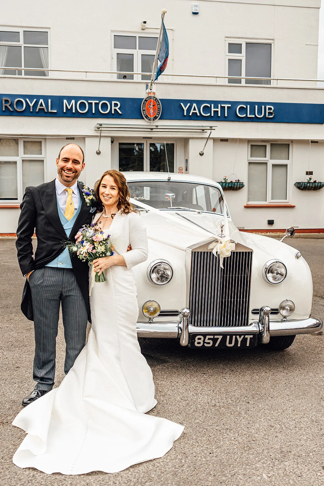 A smiling bride and groom pose in front of a classic white Rolls-Royce wedding car outside the Royal Motor Yacht Club.
