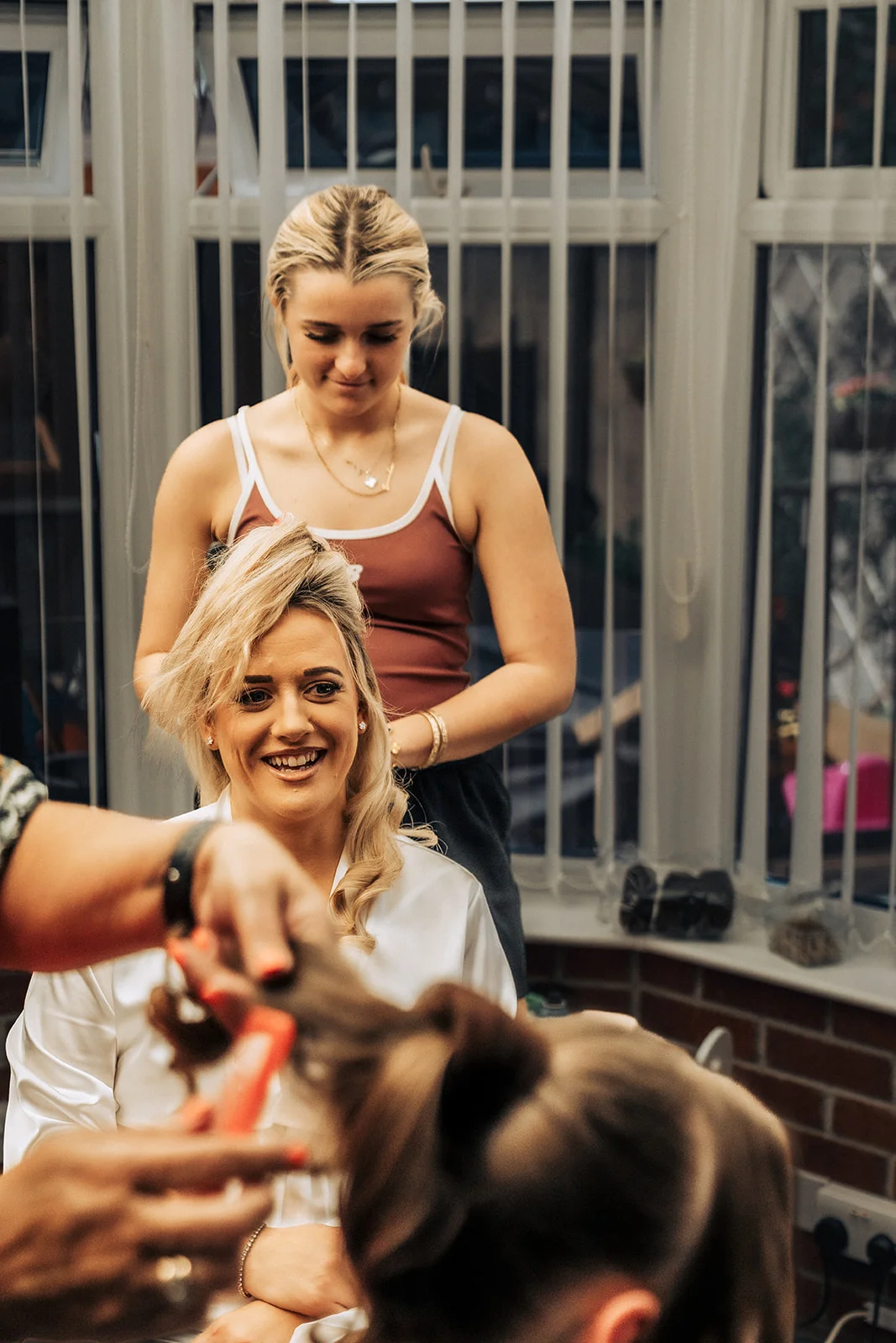 A smiling bride gets her hair styled by a bridesmaid during morning bridal preparations indoors.