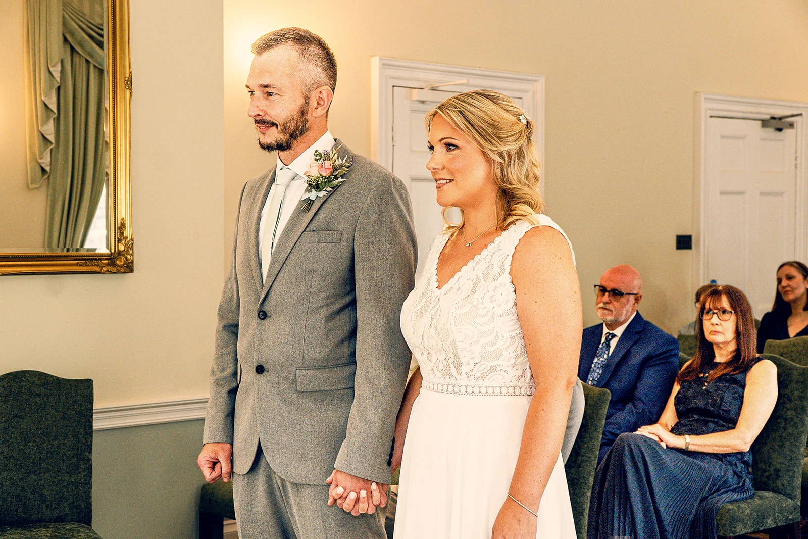 A bride in a lace wedding dress and a groom in a grey suit holding hands during a civil ceremony while guests watch from seated positions.