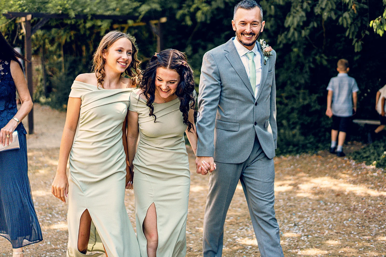 A smiling groom in a grey suit walks hand in hand with laughing bridesmaids wearing matching sage green off-the-shoulder dresses outdoors in the sunshine.
