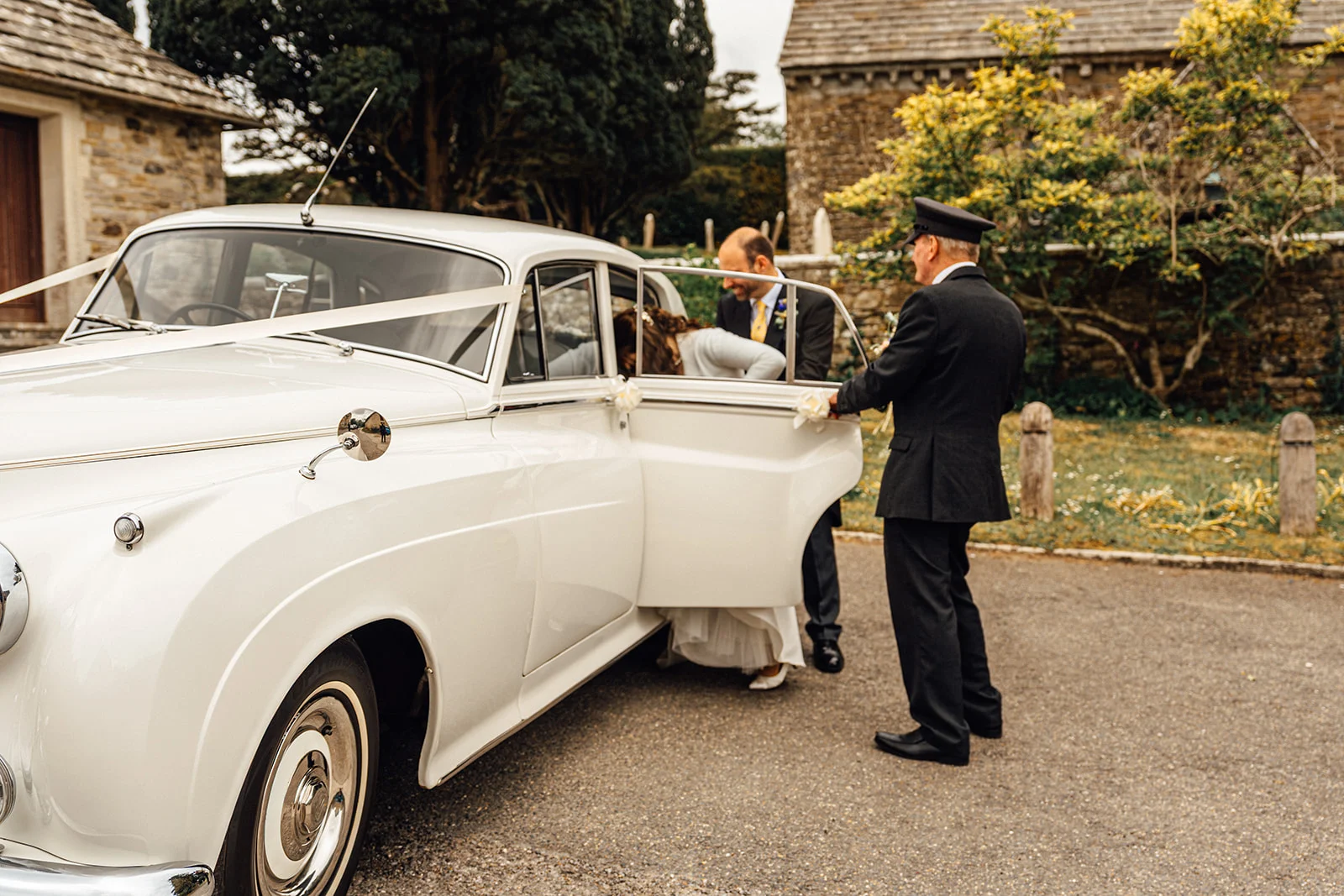 A chauffeur in uniform holds open the door of a classic white vintage wedding car adorned with ribbons as the bride arrives at the venue.