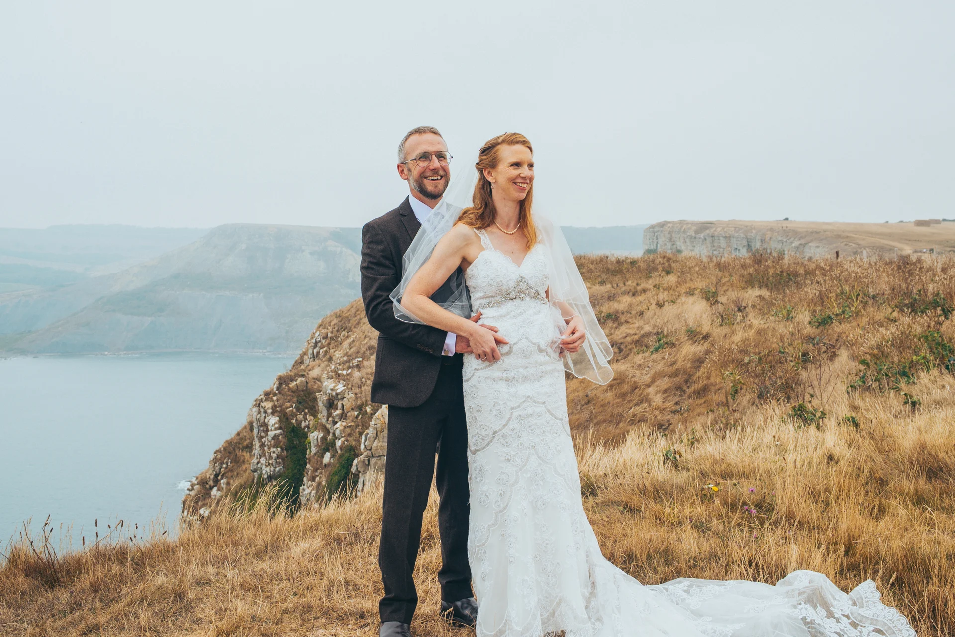 A smiling bride and groom embrace on a dramatic clifftop overlooking the sea during an overcast wedding portrait session.