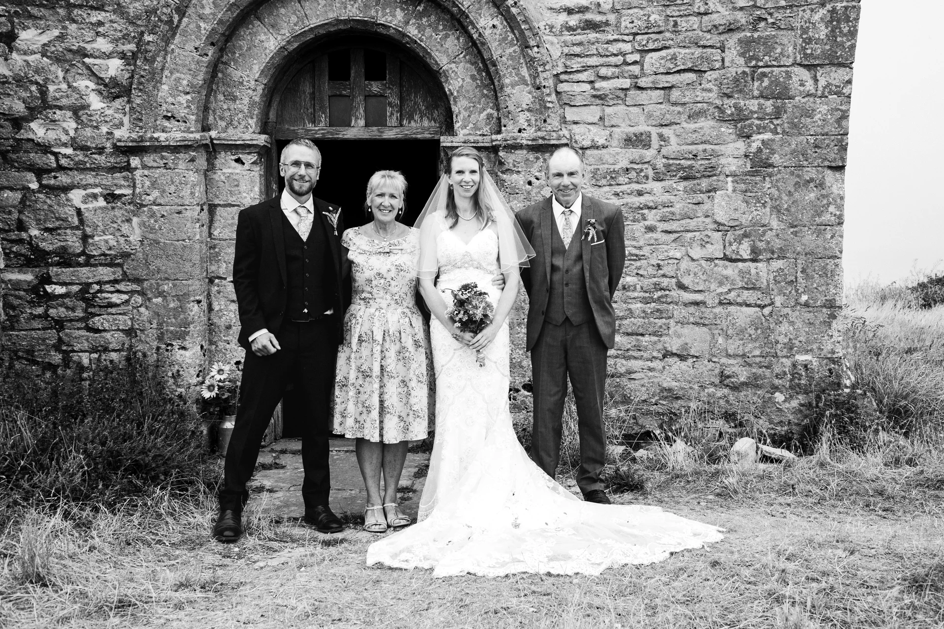 A black and white formal portrait shows the bride and groom posing outside an old stone church entrance alongside their parents.