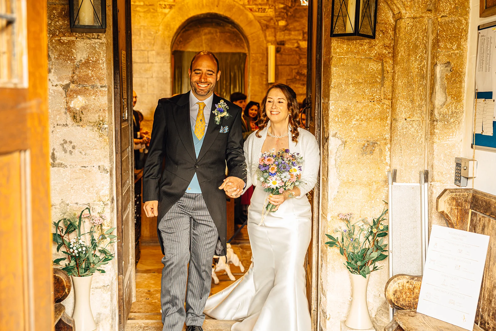 A newlywed couple smiling happily as they walk hand in hand out of a historic stone church doorway on their wedding day.