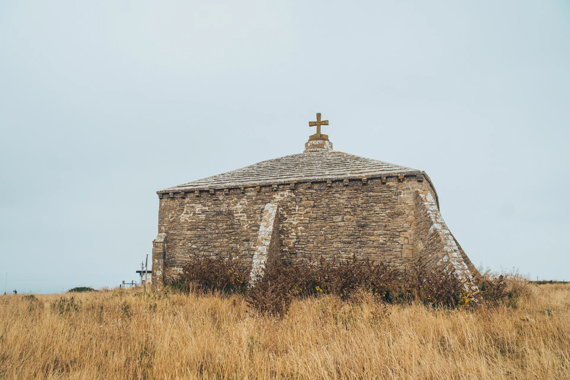 An ancient stone chapel with a rooftop cross stands solitary amid dry coastal grass under an overcast sky.