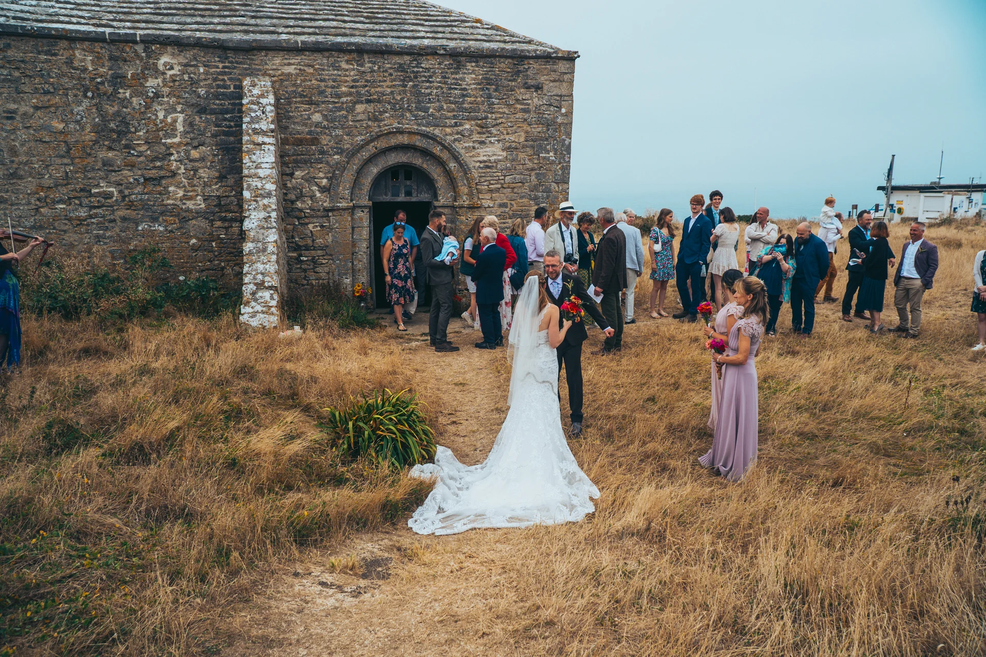 Newlyweds stand outside a rustic stone chapel on a coastal clifftop surrounded by wedding guests under an overcast sky.