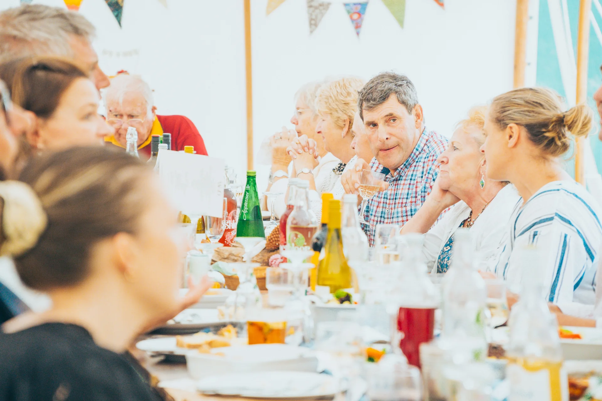 Wedding guests enjoy a relaxed wedding breakfast seated at a long banquet table inside a bright marquee decorated with bunting.