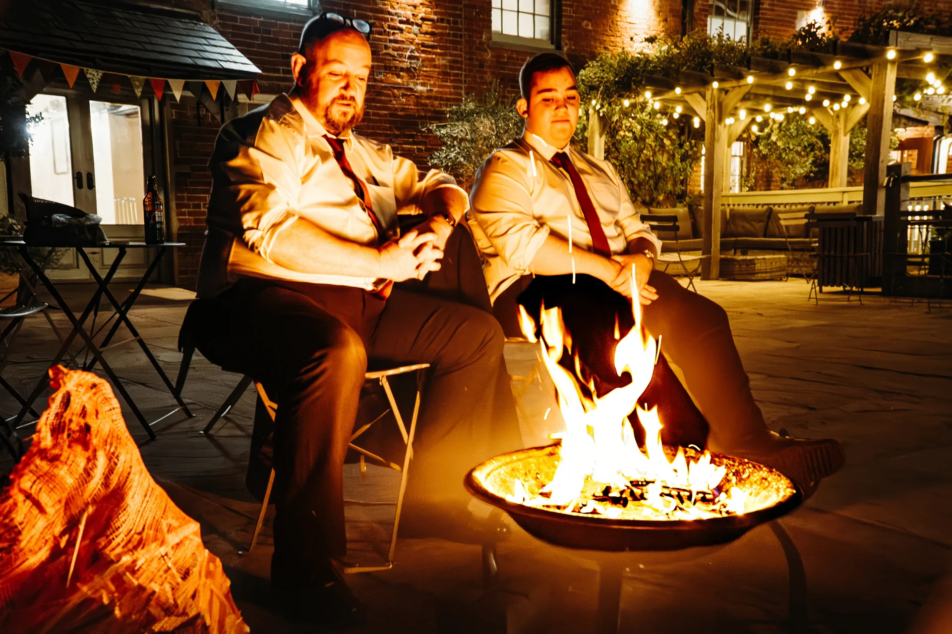 Two men seated outdoors at night by a glowing fire pit, dressed in white shirts and ties outside a brick building illuminated by festoon lights.