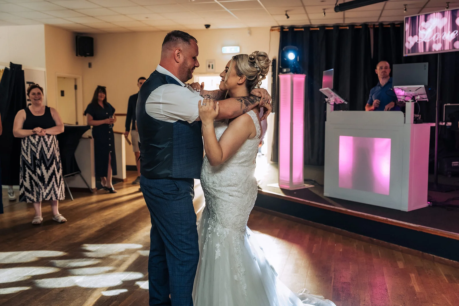 Newlyweds share their first dance together on a wooden floor in a function room, surrounded by guests and a DJ setup with pink lighting.