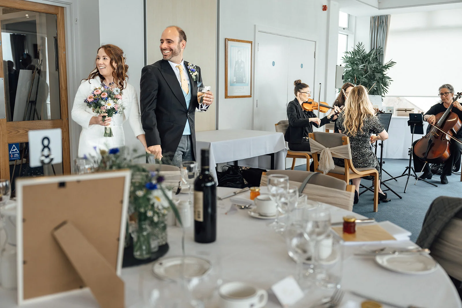 A smiling newlywed couple walk hand in hand into a reception room past tables set for the wedding breakfast and a live string trio.