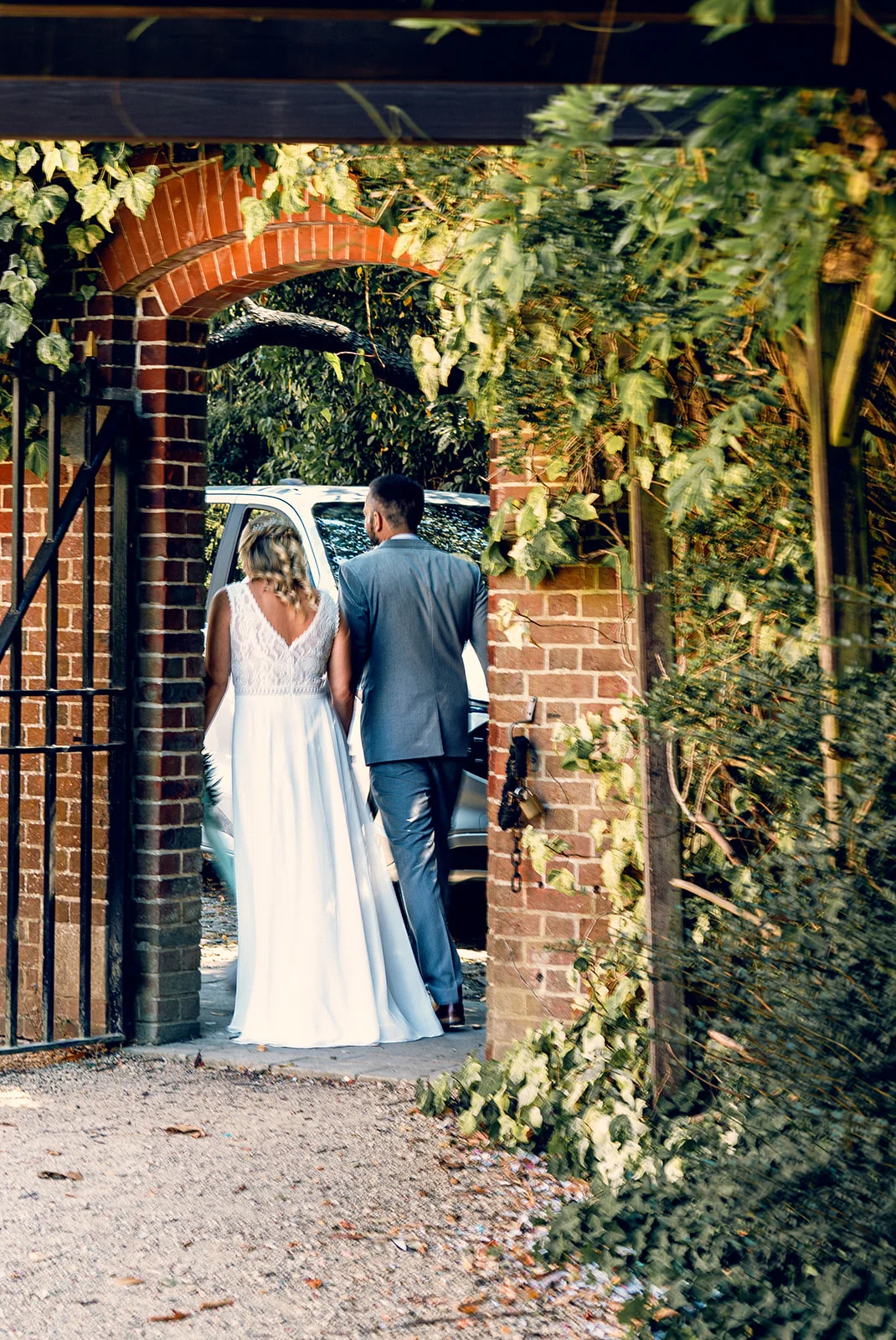 A newlywed bride and groom walk away from the camera through a brick archway towards a waiting wedding car in the warm afternoon sun.