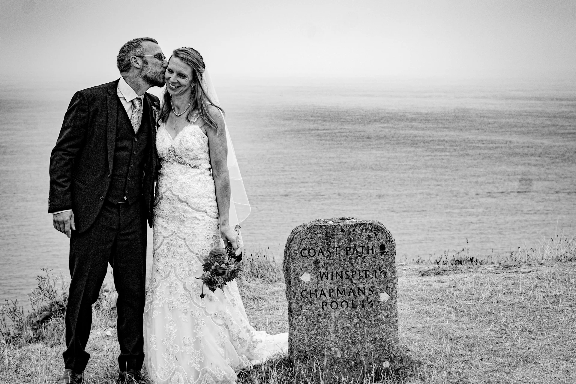 A newlywed couple poses by a coastal path sign on a clifftop, with the groom kissing the smiling bride on the cheek in a black and white photograph.