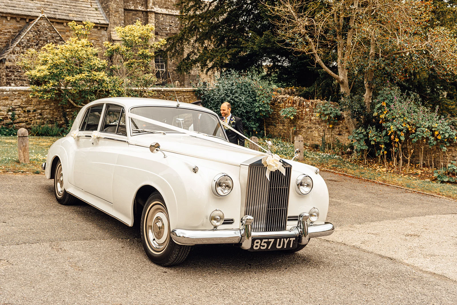 A classic white vintage wedding car decorated with ribbons is parked outside an old stone church while the groom stands nearby.