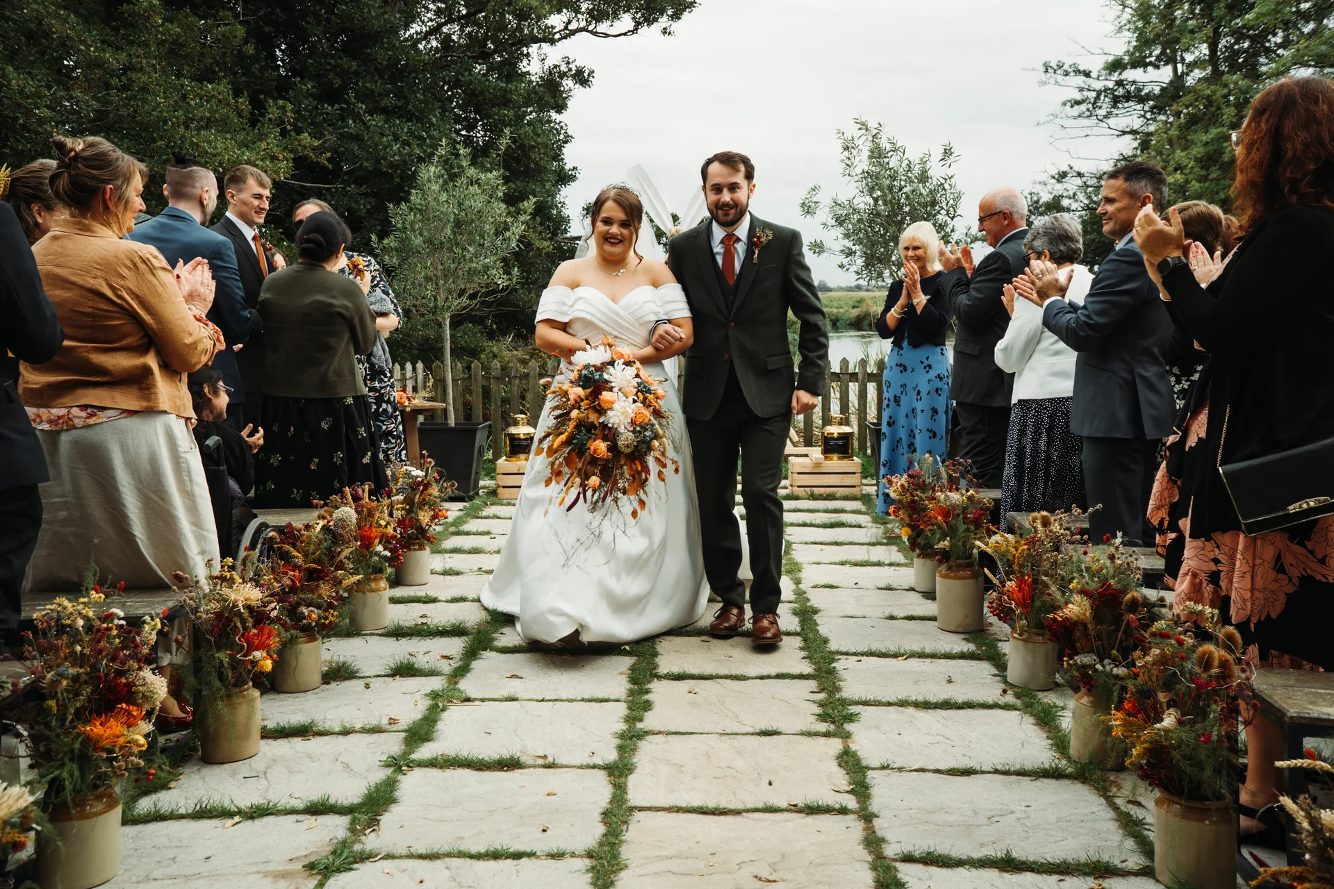 A smiling bride and groom walk hand in hand down an outdoor stone aisle lined with floral arrangements while guests applaud.