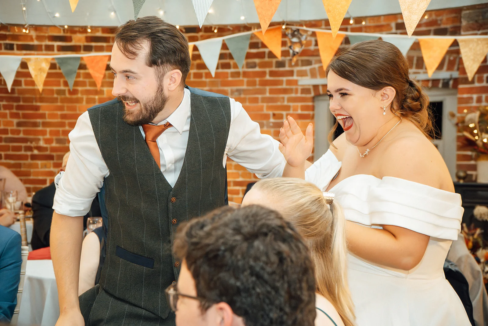 A newlywed couple laugh joyfully together during their wedding reception in a rustic barn venue decorated with orange and green bunting.