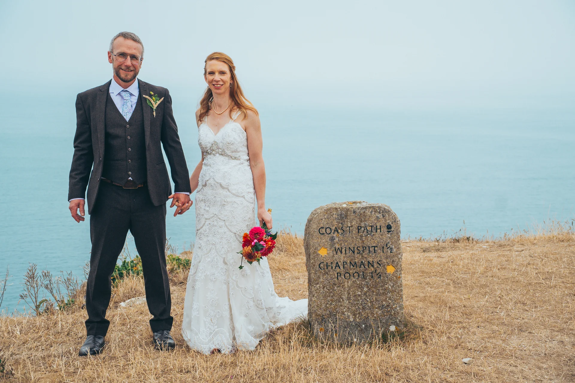 A mature bride and groom stand hand-in-hand on a coastal cliff path beside a stone signpost, with the sea stretching out behind them.