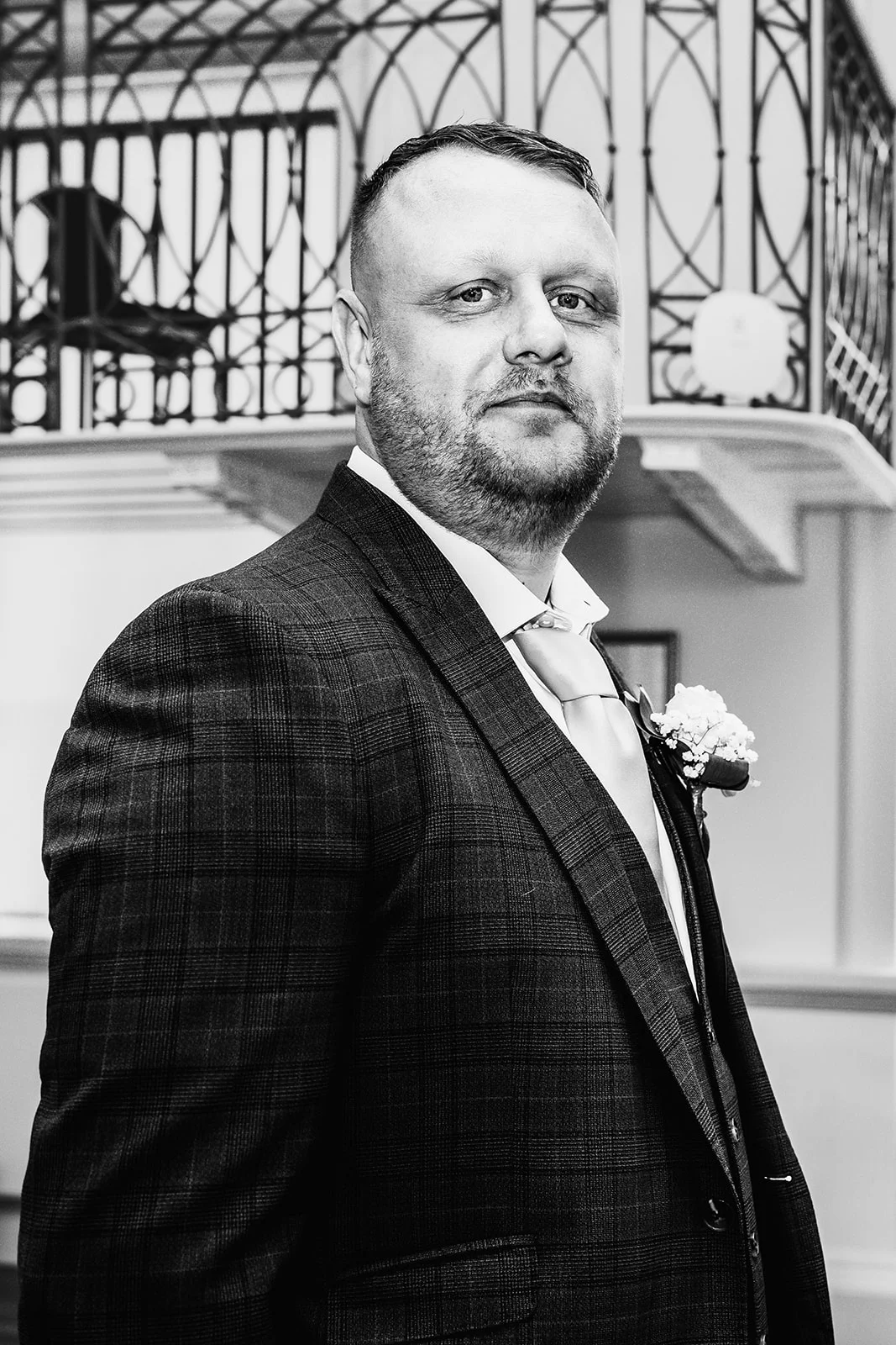 A handsome groom in a patterned checked suit stands proudly for a portrait against an indoor architectural backdrop.