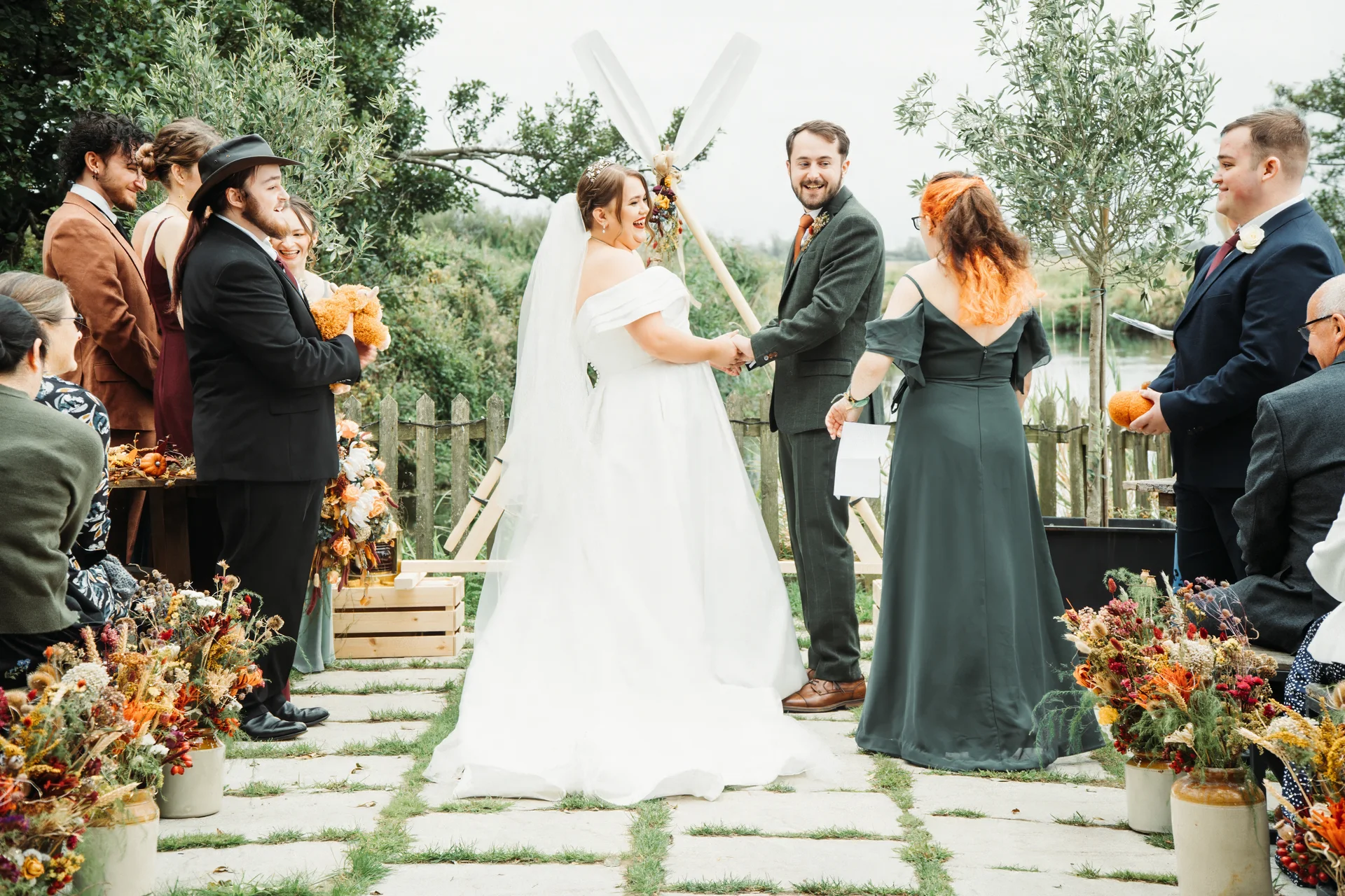 A joyful bride in an off-the-shoulder gown and veil holds hands and laughs with her groom in a tweed suit during an outdoor wedding ceremony.