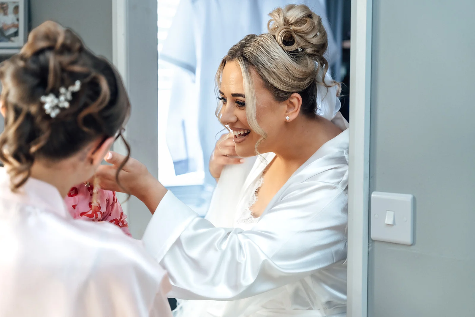 A smiling bride wearing a white satin robe and an intricate updo laughs happily while getting ready in front of a mirror with her bridesmaid.