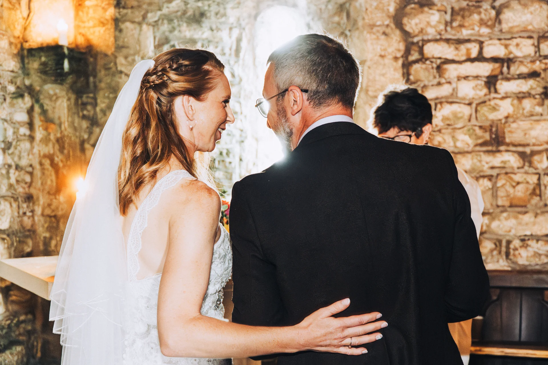 An older bride and groom share a tender, laughing embrace inside a rustic stone venue illuminated by natural backlighting.