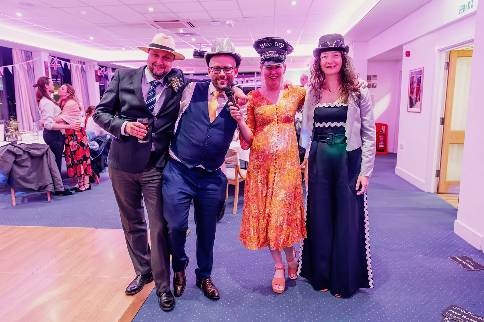 A group of wedding guests wearing novelty hats and party outfits stand together smiling under bright pink evening lighting at a reception.