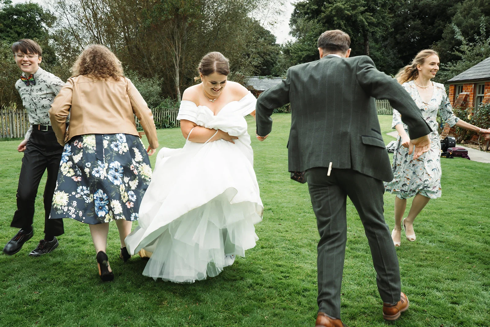 A joyful bride and groom run through a lush green garden alongside laughing wedding guests on their wedding day.
