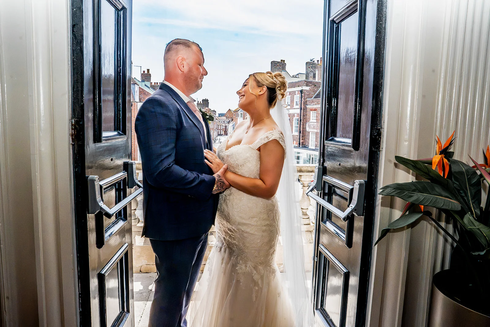 A bride and groom laughing together while framed by open double doors overlooking a city street.