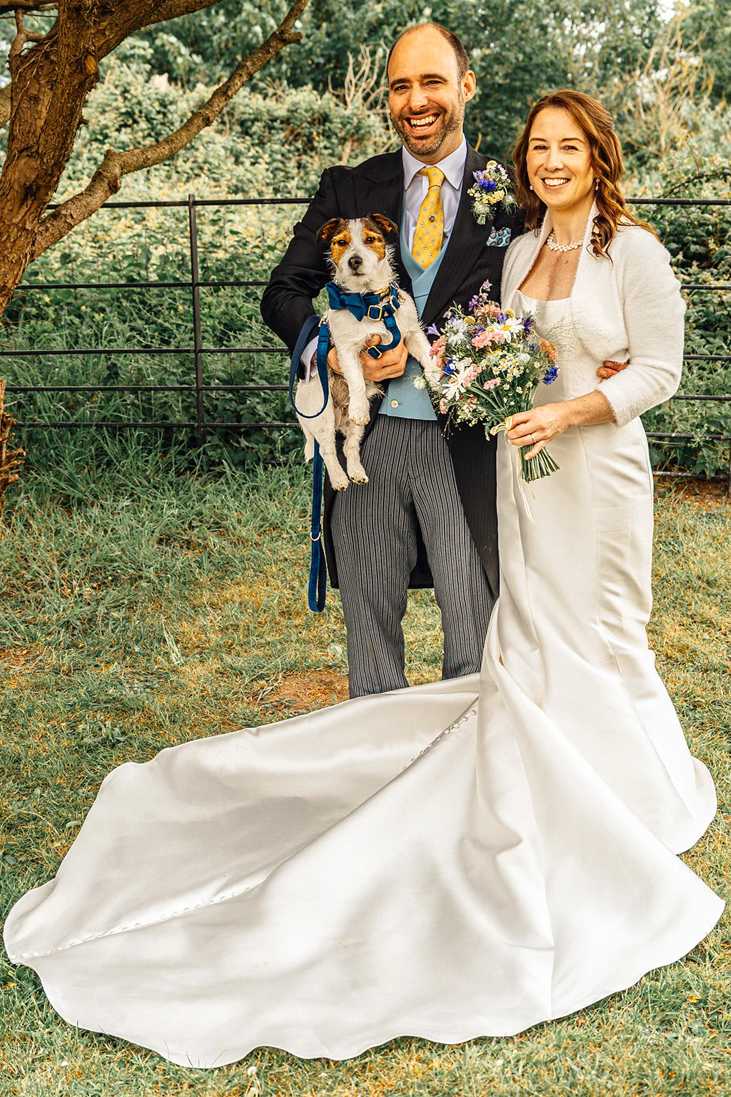 A smiling bride and groom pose outdoors in an English garden setting, holding a small Jack Russell terrier wearing a blue bow tie while the bride holds a wildflower bouquet and displays her long satin gown with a dramatic train.
