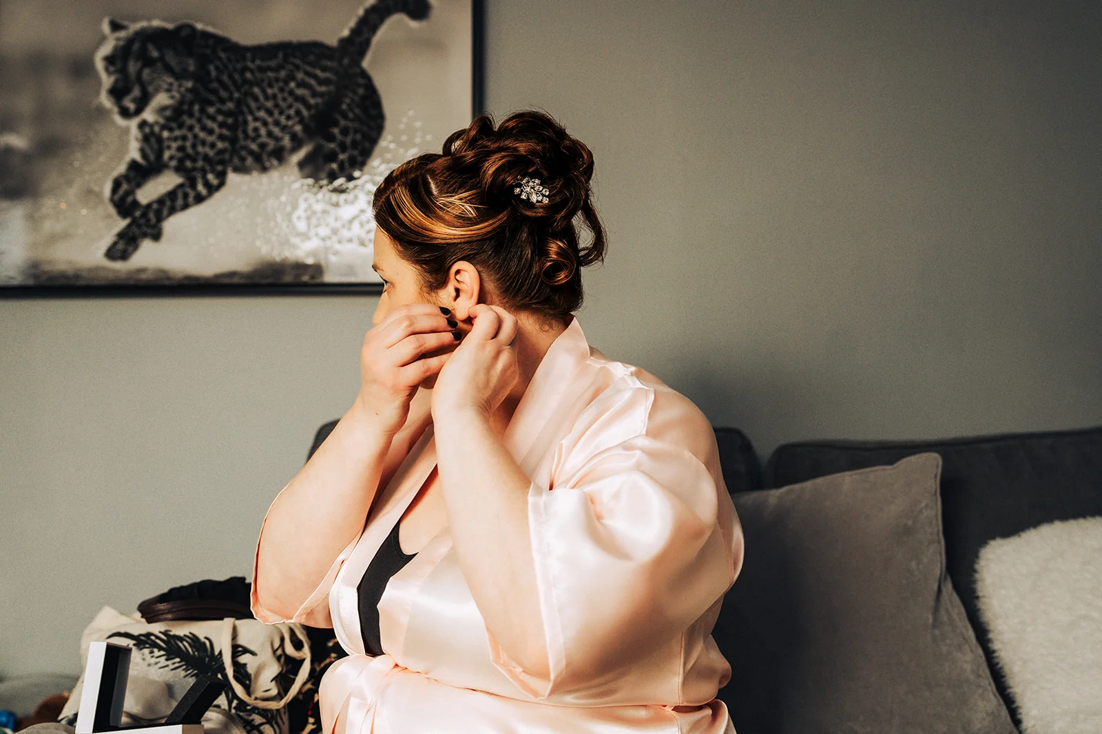 A bride in a pink satin robe sits indoors putting on her earrings while her hair is styled in an intricate updo with a decorative hair comb.