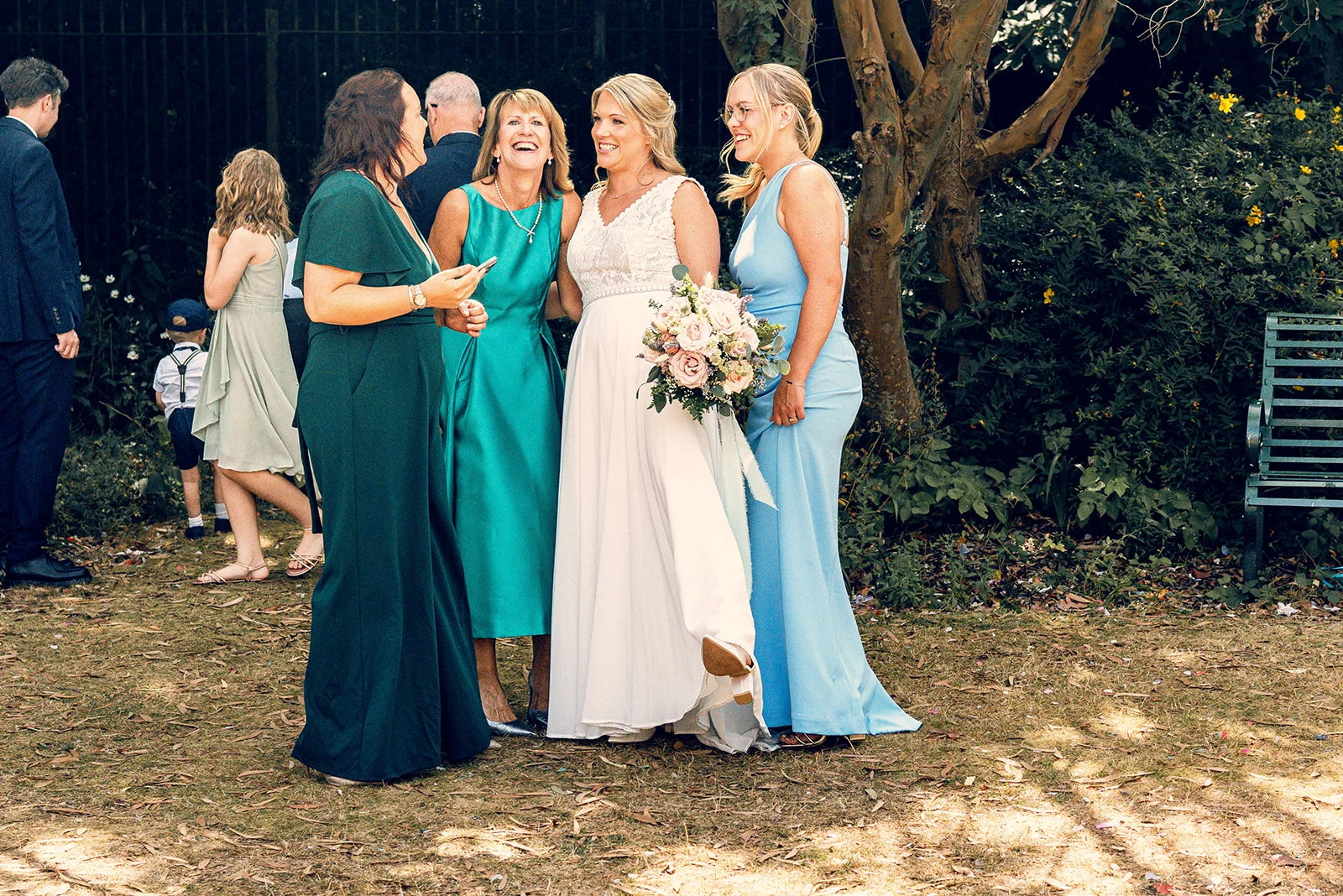 A smiling bride holding a bouquet stands outdoors in a garden setting surrounded by laughing family members and bridesmaids.