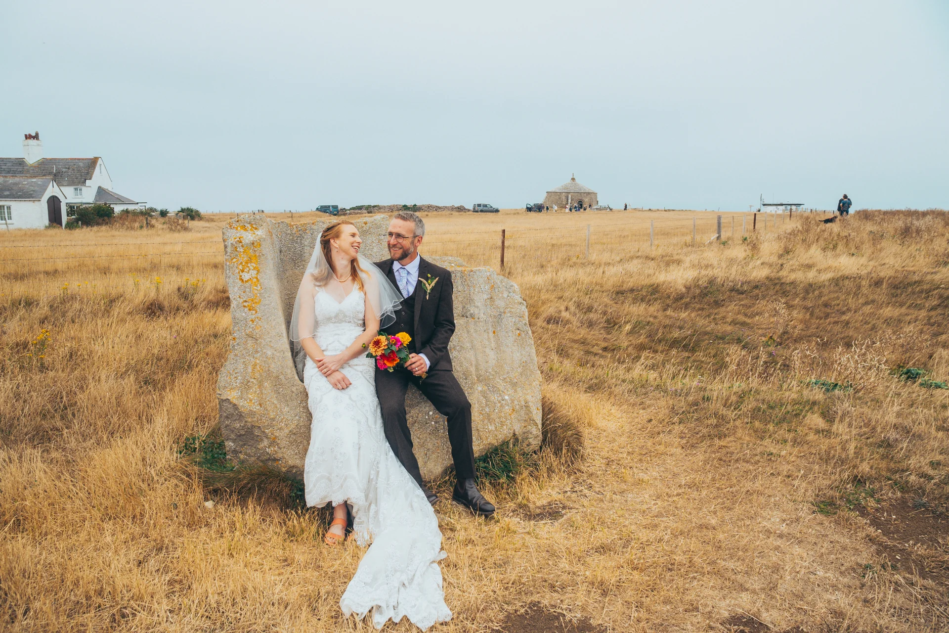 A newlywed couple sits smiling together on a large stone sculpture in the middle of a vast, grassy coastal landscape under an overcast sky.