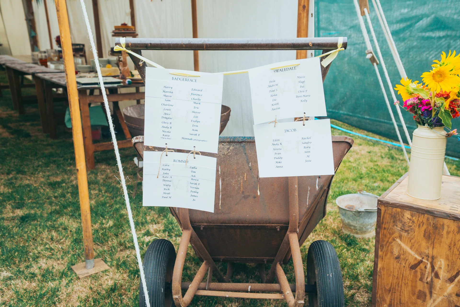 A rustic wedding seating plan is creatively displayed on an old rusty wheelbarrow outside a marquee, accompanied by a vibrant arrangement of sunflowers.