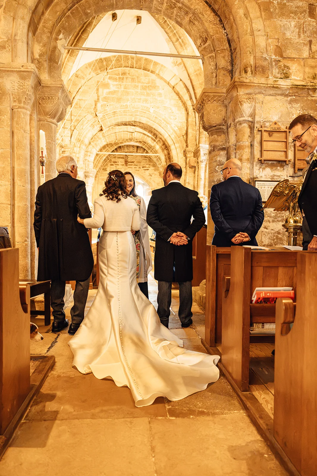 The bride walks down the aisle of an ancient stone church on her father's arm, approaching the groom and wedding party standing at the altar.