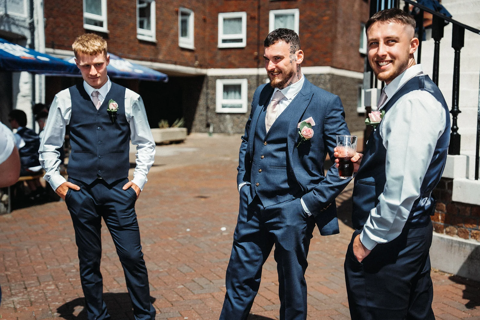 Three smiling groomsmen in matching navy suits and waistcoats stand outside a brick building on a sunny day during the wedding reception.