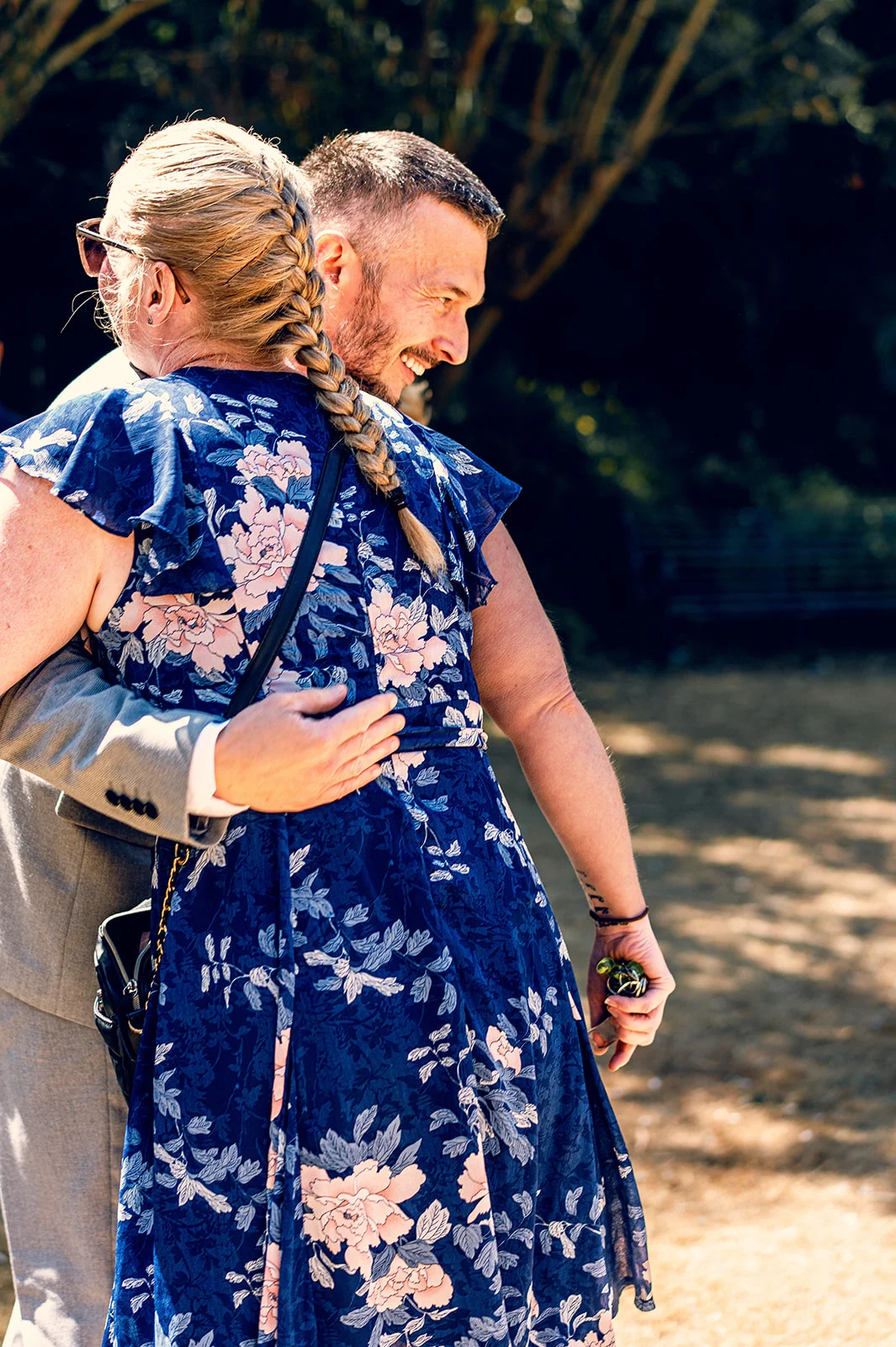 A guest in a blue floral dress embraces a smiling groom wearing a grey suit during an outdoor wedding celebration.