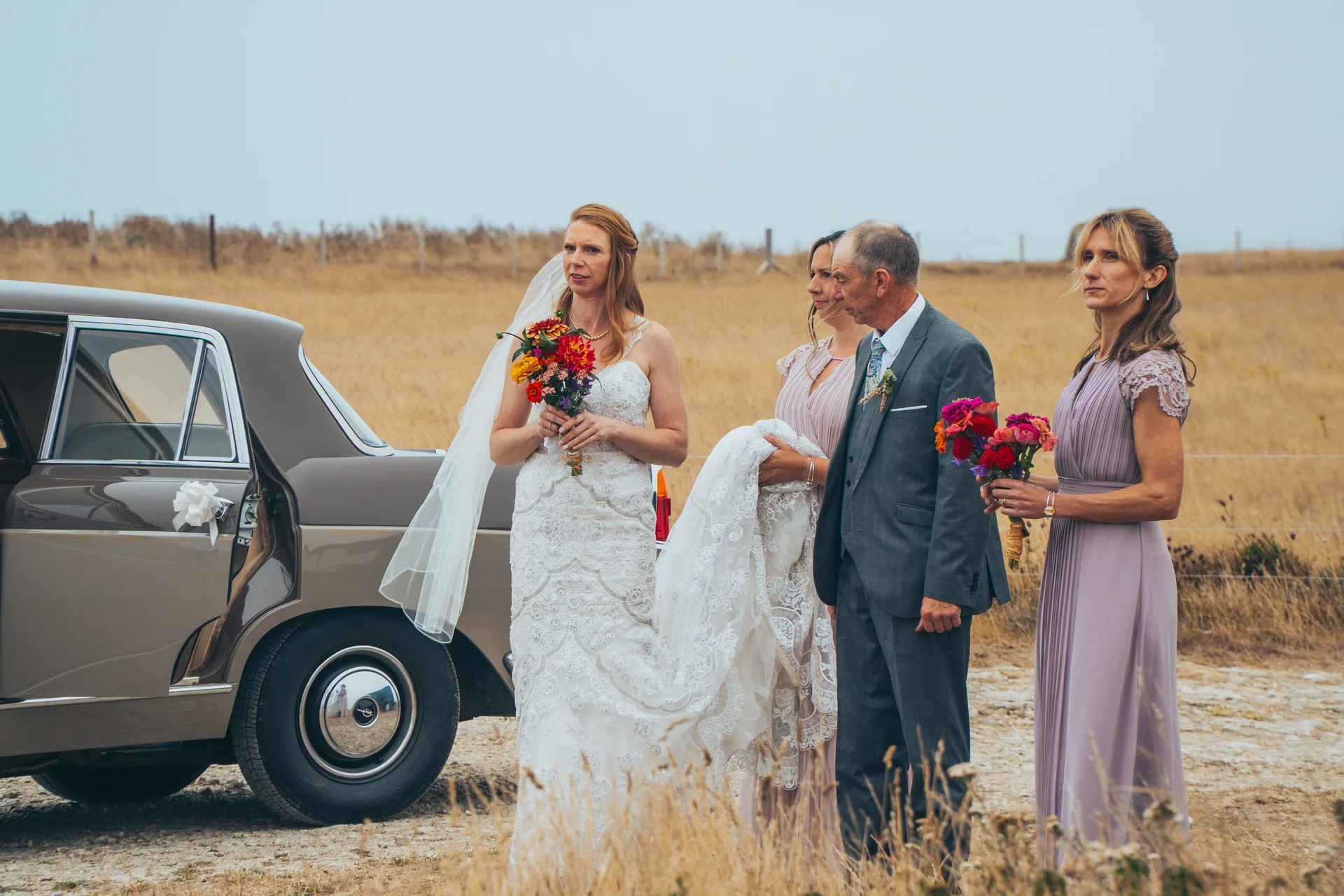 A bride holding a colourful bouquet stands next to a classic car alongside her father and bridesmaids in an outdoor rural setting.