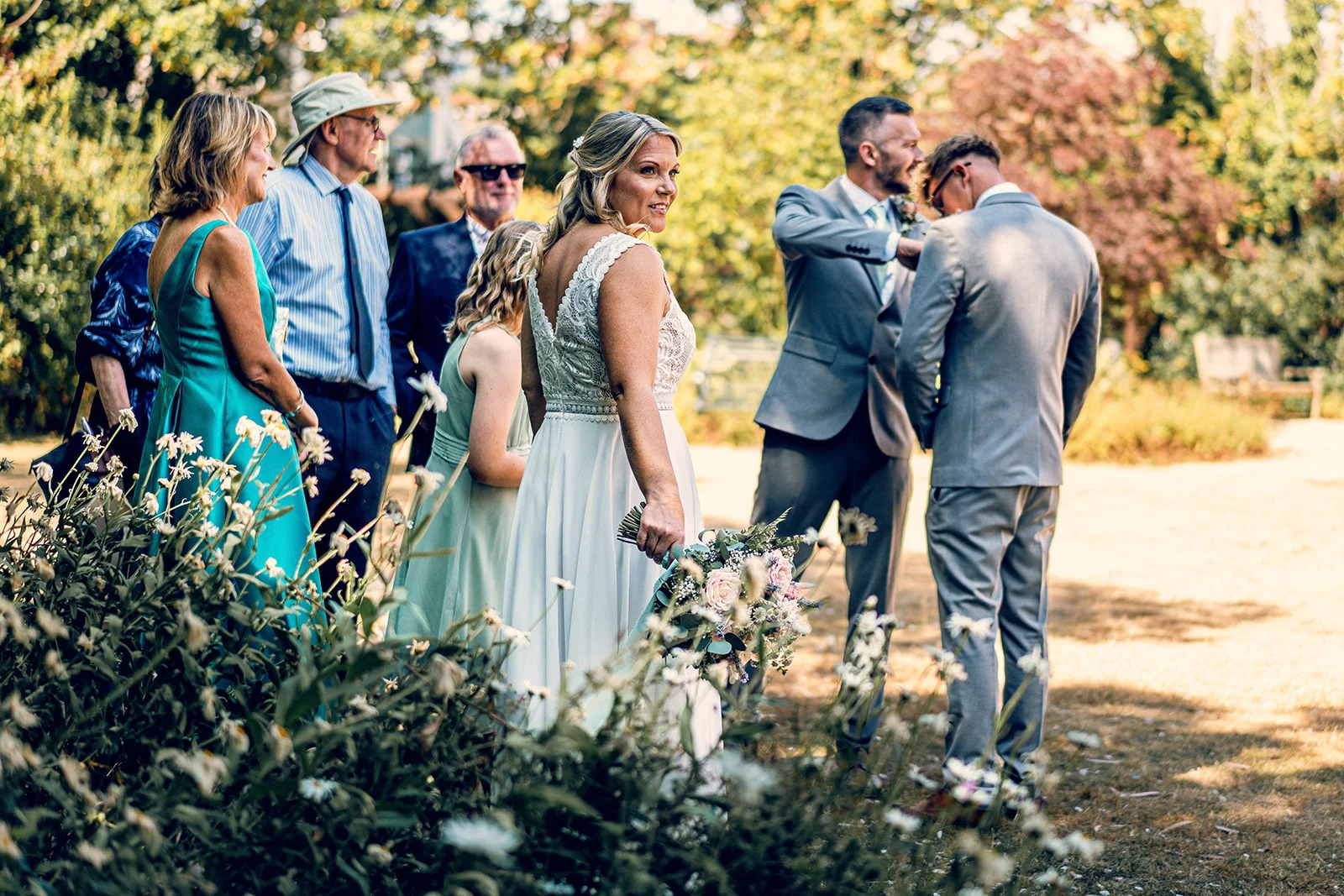 A smiling bride holding a floral bouquet glances back over her shoulder outdoors, framed by garden foliage while wedding guests and groomsmen stand nearby in the warm sunshine.