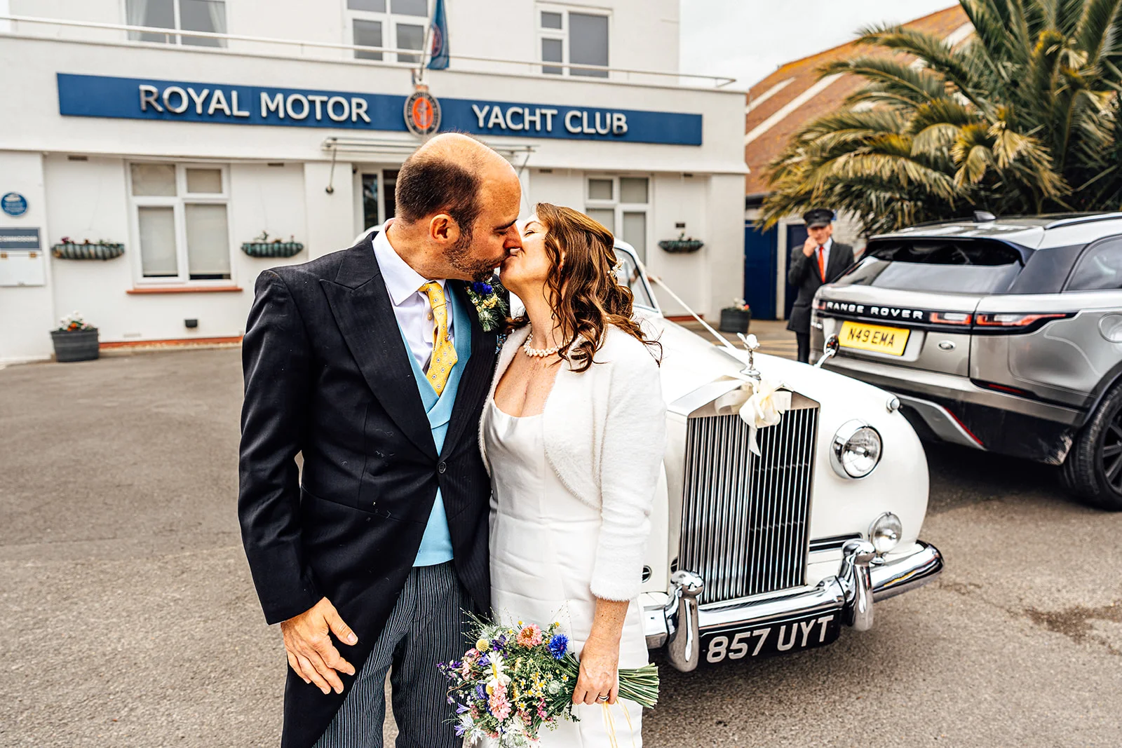 Newlywed bride and groom share a kiss outside a yacht club in front of a classic white vintage wedding car.