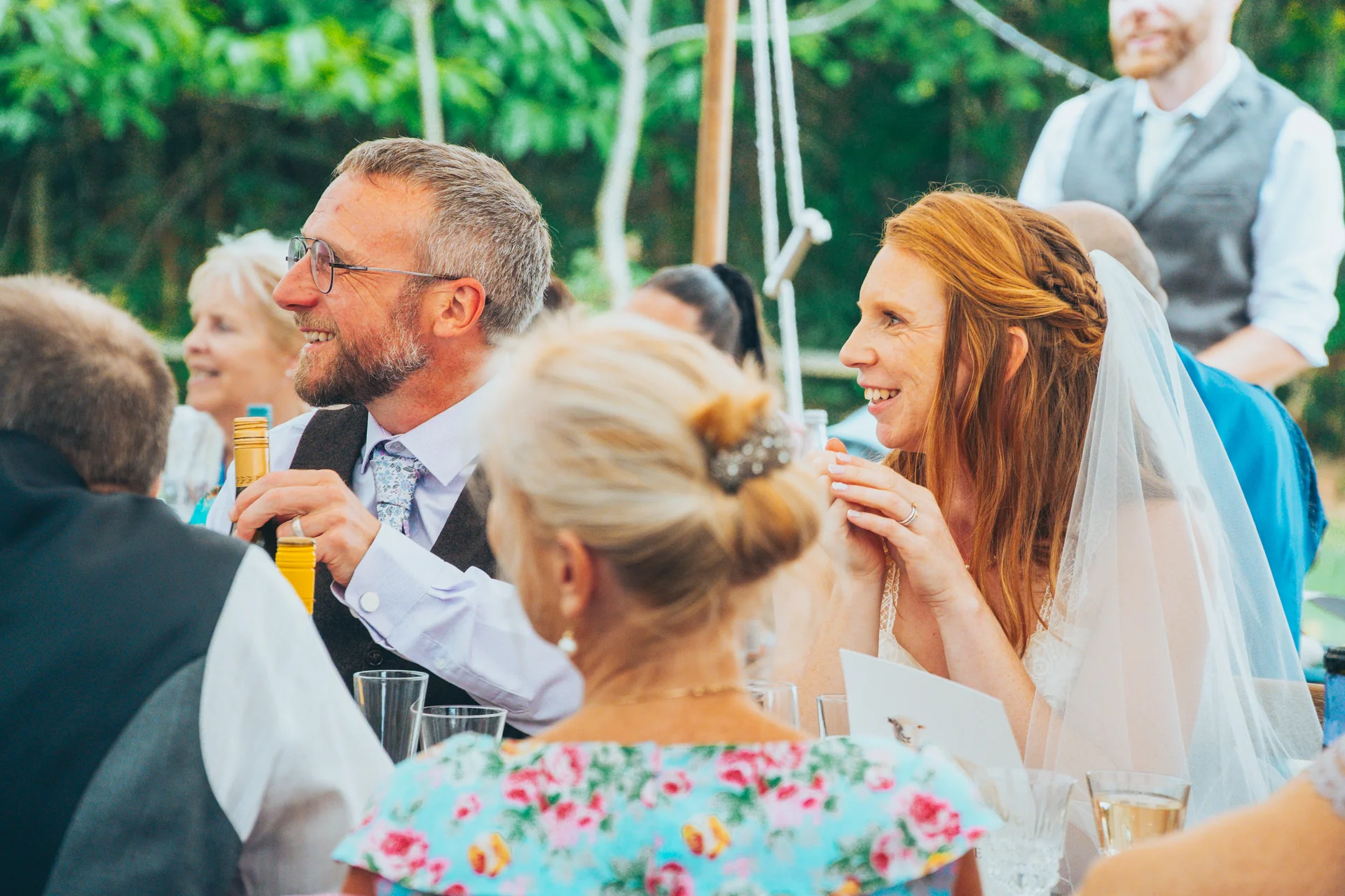 The bride and groom laugh heartily during the wedding breakfast inside a marquee, surrounded by smiling guests and family members.
