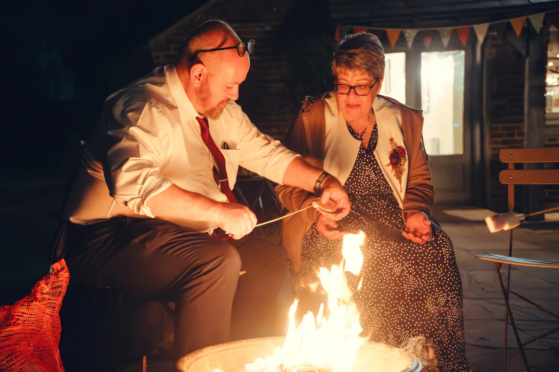 An older couple enjoys toasting marshmallows together over a glowing firepit outdoors at night during a wedding reception.