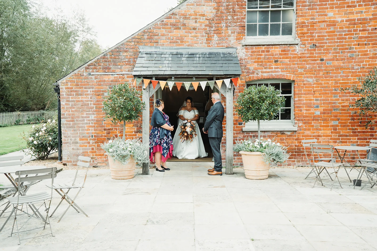 A bride holding a bouquet stands under a porch of a rustic red brick barn with guests gathered outside for a wedding ceremony.
