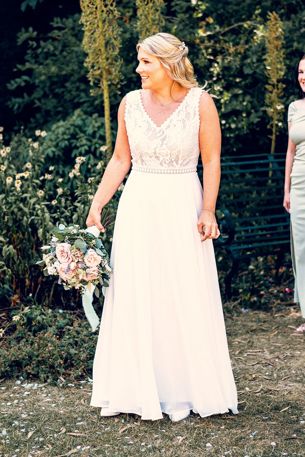 A smiling bride holding a hand tied rose bouquet stands in a garden wearing an a line lace dress and half up hairstyle.